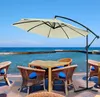 An outdoor patio setting with a large beige cantilever umbrella providing shade over a table and chairs, with a body of water and a clear blue sky in the background. Voir les images du produit