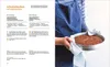 A person is holding a round cake in a baking pan, with a recipe book open next to it. Produktbild-Vorschau