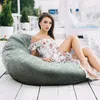 A woman is sitting on a large bean bag chair on a wooden deck, with trees and white structures in the background. Náhled obrázku produktu
