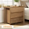 A wooden nightstand with three drawers is placed next to a bed, with a window in the background. Produktbild-Vorschau