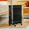 A black oil-filled radiator is placed on a woven rug next to a bed and a wicker chair in a room. Produktbild-Vorschau