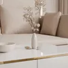 A close-up of a white coffee table with a vase of dried flowers and a marble bowl on it, with a beige sofa in the background. Produktbild-Vorschau