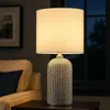 A table lamp with a white shade and a textured silver base is placed on a dark wooden side table next to a cream-colored armchair with a dark blue pillow. Anteprima dell’immagine prodotto