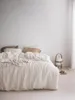 A neatly made bed with white linen bedding, pillows, and a throw blanket. A bedside lamp and slippers are visible to the right. Produktbild-Vorschau