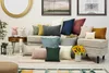 A collection of decorative throw pillows arranged on a sofa and ottoman, with decorative items like a vase of sunflowers, a mirror, and framed art in the background. Produktbild-Vorschau