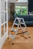A step ladder is placed on a wooden floor in a living room, near a doorway. A sofa and curtains are visible in the background. Produktbild-Vorschau