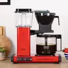 A red Moccamaster coffee maker is shown on a countertop with a strawberry in a bowl in the background. Náhled obrázku produktu