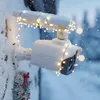 A white security camera is mounted on a snowy wall, decorated with string lights and a small wreath. Produktbild-Vorschau