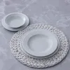 A set of white porcelain plates and bowls are arranged on a textured placemat on a marble-like surface. Náhľad obrázku produktu