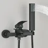 A black bathtub faucet with a showerhead attached, mounted on a light gray wall. Water is spraying from the showerhead. Náhled obrázku produktu