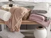 A collection of soft blankets and pillows arranged on a wooden bench, with a tray of coffee and a book in the background. Náhled obrázku produktu