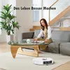A woman is sitting on a sofa reading a magazine while a robot vacuum cleaner operates on the floor. Produktbild-Vorschau