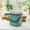 A teal compost bin with a gray lid and handle sits on a white countertop in a kitchen setting. Various vegetables and plants are visible in the background. Produktbild-Vorschau