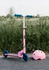 A pink scooter and a pink helmet are parked on a paved surface next to tall green grass. Produktbild-Vorschau
