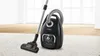A black Bosch vacuum cleaner with a silver hose and wand is placed on a white carpet, with a light wood floor visible in the background. Produktbild-Vorschau
