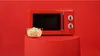 A red retro-style microwave oven is displayed on a red cylindrical stand against a red background. A bowl of popcorn is placed in front of the microwave. Anteprima dell’immagine prodotto