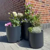 Three dark gray planters filled with various flowers and greenery are placed on a paved patio in front of a brick wall. Podgląd zdjęcia produktu