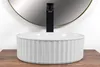 A white ceramic washbasin with a fluted exterior sits on a wooden surface in front of a large round mirror and a black faucet. Produktbild-Vorschau