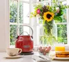 A kitchen counter with a red kettle, a cup of tea, a bowl of strawberries, and a sandwich, placed in front of a window with a bouquet of flowers. Produktbild-Vorschau