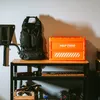 A black backpack and an orange storage crate are displayed on a wooden shelf against a light gray wall. Produktbild-Vorschau