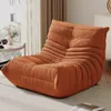 A modern, plush orange floor sofa is shown in a well-lit living room setting, next to a window and a small side table. Náhľad obrázku produktu