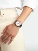 A close-up shot of a woman's wrist wearing a rose gold watch with a black band, against a neutral background. Produktbild-Vorschau