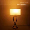 A table lamp with a rectangular shade is placed on a reflective surface against a gradient brown background. Produktbild-Vorschau