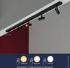 A black track light fixture mounted on a ceiling, with a dark red wall in the background. The bottom of the image shows three different light intensity settings. Náhľad obrázku produktu