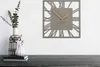 A modern wall clock with Roman numerals is displayed on a white wall above a dark wooden surface with two vases and books. Náhled obrázku produktu