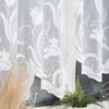 A close-up view of a white lace curtain with a floral pattern, partially obscuring a window with a dark, marbled-patterned blind and a potted fern. Produktbild-Vorschau