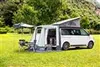 A white camper van with a pop-top roof and an attached tent is parked on a grassy field with trees in the background. Produktbild-Vorschau