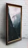 A framed picture of a misty mountain landscape is leaning against a white brick wall. Produktbild-Vorschau