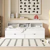 A white daybed with storage drawers and a bookshelf headboard, set in a minimalist bedroom with a patterned rug and artwork on the wall. Náhled obrázku produktu