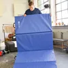 A man is holding a large blue folding mat in a room with large windows. Produktbild-Vorschau