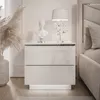 A white, glossy nightstand with two drawers, a bust sculpture, and a decorative lamp on top, placed in a bedroom setting. Produktbild-Vorschau