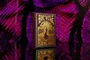 A deck of Bicycle playing cards with a peacock design, placed on a reflective surface with a vibrant purple and black abstract background. Produktbild-Vorschau