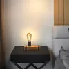 A vintage-style table lamp with a wooden base sits on a black nightstand next to a bed. Produktbild-Vorschau