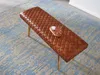 A brown leather bench with a quilted pattern is placed on a blue and beige rug. A small wooden tray with a drink and a small bowl is on the bench. Produktbild-Vorschau