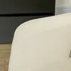 A close-up view of the armrest and back of a beige upholstered chair, with a dark cabinet and wooden floor visible in the background. Anteprima dell’immagine prodotto