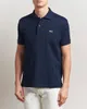 A man wearing a navy blue Lacoste polo shirt and white pants, shown from the chest down against a white background. Produktbild-Vorschau