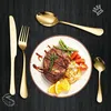 A gold cutlery set is arranged around a plate of food, including steak, pasta, and vegetables, on a black wooden table. Anteprima dell’immagine prodotto