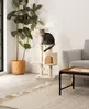A cat is sitting on a beige cat tree next to a large potted plant and a gray sofa. Produktbild-Vorschau