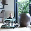 A baby is sitting in a car-shaped baby walker in a room with a large plant and a floor lamp. Produktbild-Vorschau