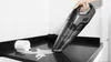 A hand is holding a black and gray handheld vacuum cleaner over a black countertop, cleaning up a spill from a white mug. Podgląd zdjęcia produktu
