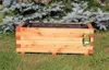 A wooden planter box with a black liner sits on green grass in front of a blurred background of evergreen trees. Produktbild-Vorschau