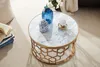 A round coffee table with a marble top and a gold geometric base, decorated with a blue vase and a decorative jar. Produktbild-Vorschau