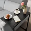 A black C-shaped side table with a croissant, coffee cup, notebook, and a rose in a vase on it, placed next to a light gray sofa. Produktbild-Vorschau