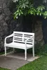 A white bench sits on a concrete path in a garden, with a stone wall and foliage in the background. Produktbild-Vorschau