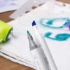 Close-up of two dual-tip markers on a white surface with some stencil art and a green airbrush gun in the background. Produktbild-Vorschau