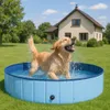 A golden retriever dog is playing and splashing in a blue, round, foldable dog pool in a grassy backyard with a house in the background. Produktbild-Vorschau